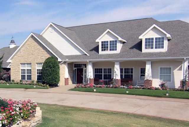 Front exterior view of a single-story residential building with a gabled roof, multiple windows, a front porch with columns, and landscaped flower beds and bushes along the walkway and lawn.