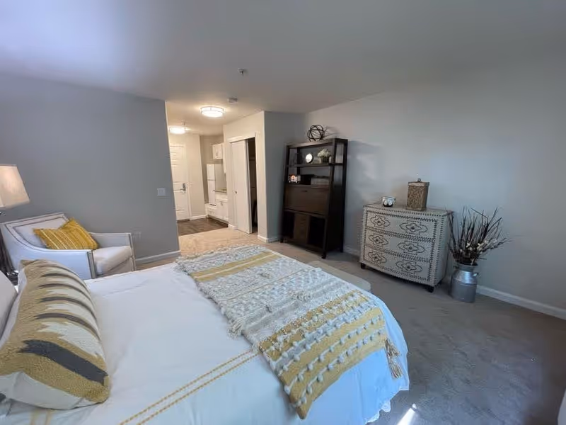 A bedroom with a neatly made bed featuring a white and yellow patterned blanket and pillows. To the left of the bed is a white armchair with a yellow pillow and a floor lamp. In the background, there is a small kitchenette area with white cabinets and a refrigerator. On the right side of the room, there is a black shelving unit with decorative items and a white dresser with a decorative box on top. A large vase with dried branches is placed on the floor next to the dresser. The walls are light-colored and the room is well-lit with natural light.