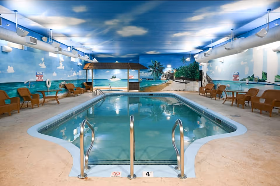 Indoor swimming pool with a tropical beach mural painted on the walls and ceiling. The pool area has several wicker chairs and glass-top tables along the sides. The ceiling is painted blue with white clouds, and there are metal handrails at the pool entrance.