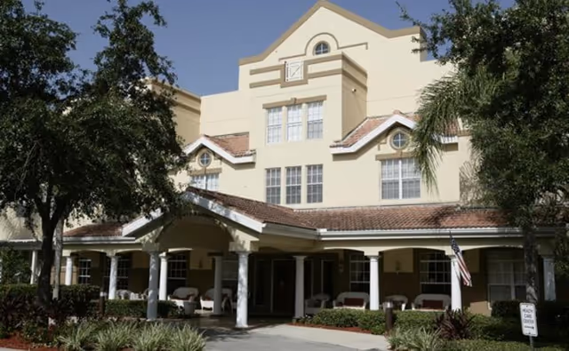 Front exterior view of a multi-story senior living facility building with a covered entrance supported by white columns, surrounded by trees and landscaping, with several chairs on the porch and an American flag displayed.
