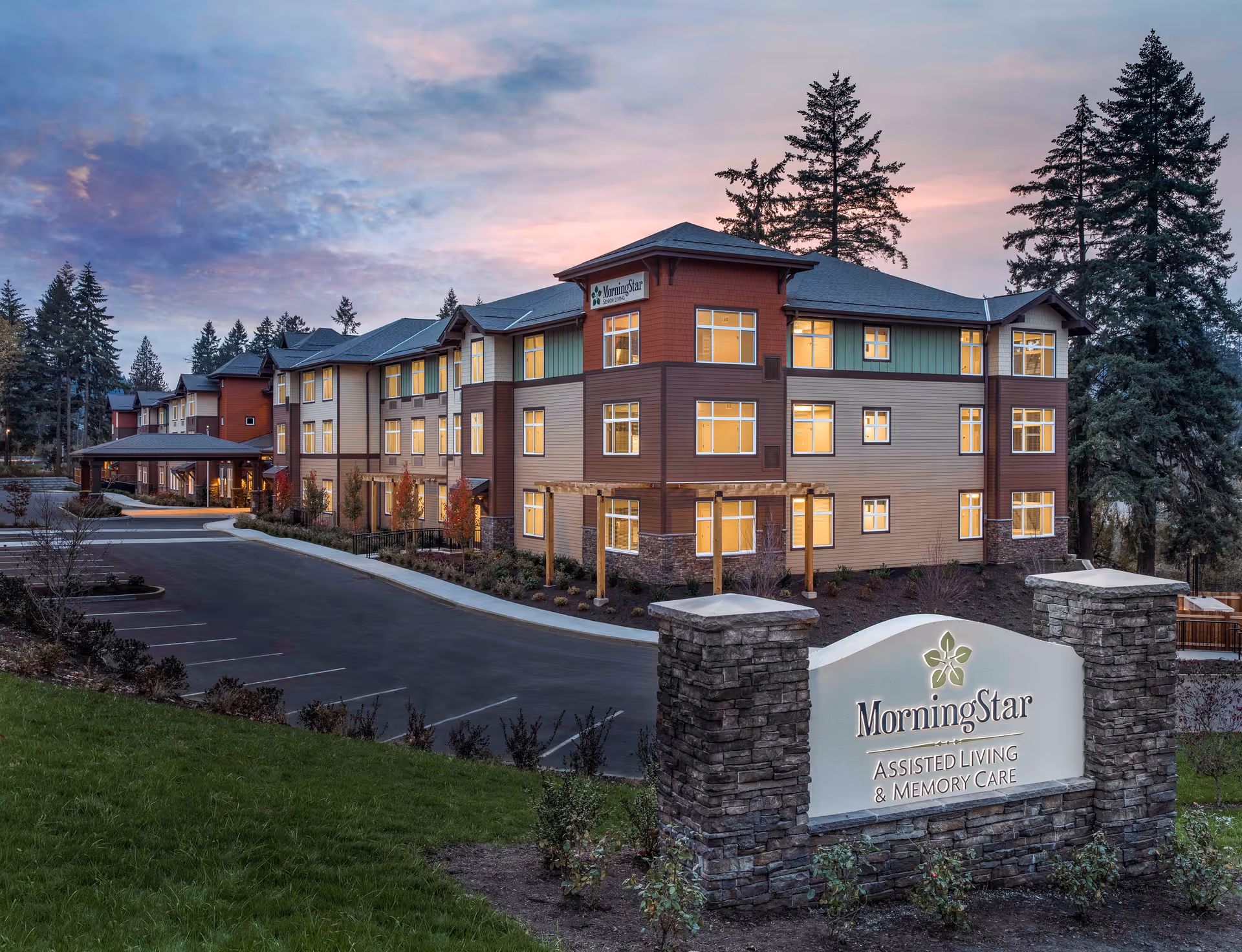 Exterior view of the MorningStar Assisted Living & Memory Care facility at dusk, showing a three-story building with lit windows, surrounded by trees and a parking area. A stone sign with the facility's name is visible in the foreground.