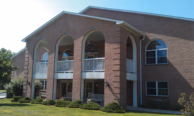 Exterior view of a two-story brick building with arched openings on the upper balcony and large windows. The building is surrounded by green grass, shrubs, and trees under a clear blue sky.
