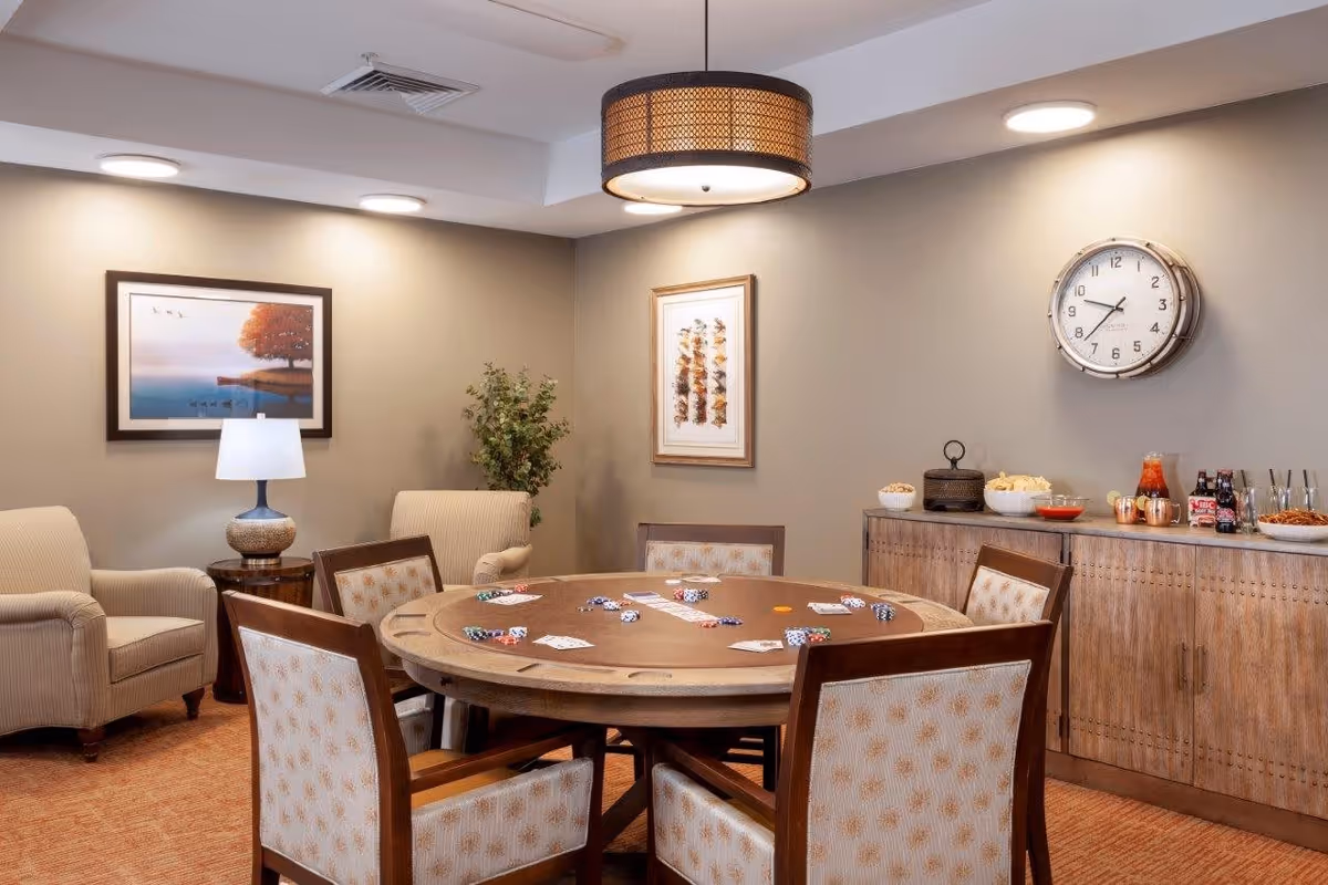 A cozy room with a round poker table surrounded by six cushioned chairs. The table has poker chips and playing cards scattered on it. In the background, there are two beige armchairs with a side table and lamp between them, a plant, framed artwork on the walls, a wall clock, and a wooden cabinet with snacks and drinks on top. The room is softly lit with ceiling lights and a hanging lamp above the table.