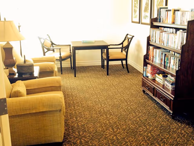 A cozy room with two beige armchairs, a small table with a lamp, a wooden table with two chairs, and a wooden bookshelf filled with books and board games. The room has patterned carpet and framed pictures on the wall.