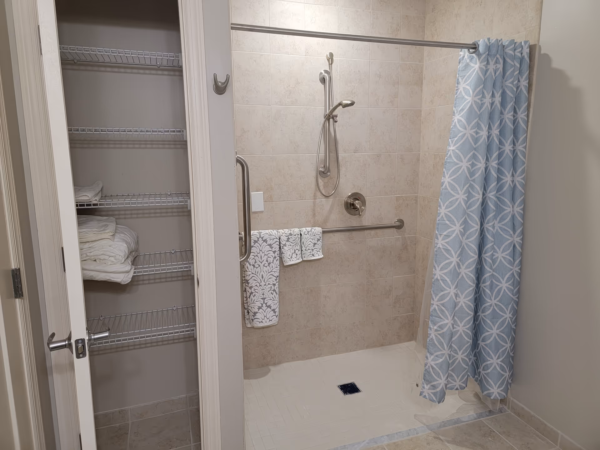 A tiled walk-in shower with a blue and white patterned shower curtain, a handheld showerhead, and grab bars. Next to the shower is an open linen closet with wire shelves holding folded towels and linens.