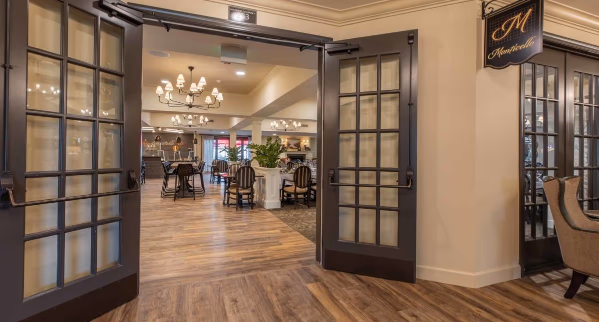 View through open double glass doors into a spacious dining room with wooden floors, multiple round tables with chairs, chandeliers hanging from the ceiling, and plants placed on a white column in the center. A sign with the word 'Monticello' is visible on the right wall.