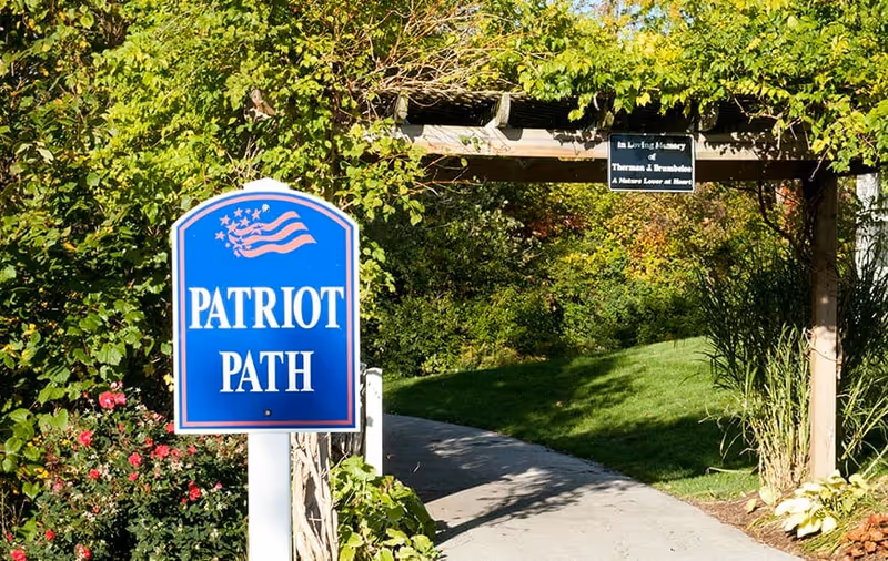 A paved garden path named Patriot Path with a blue sign featuring a waving American flag design. The path is surrounded by green trees, bushes, and flowers, and there is a wooden pergola overhead with a small memorial plaque.