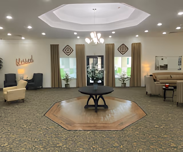 A spacious, well-lit common area with a central round table on a wooden octagonal platform. The room has patterned carpet flooring, beige walls, and a white ceiling with recessed lighting and a chandelier. There are large windows with beige curtains, two armchairs with a small table and lamp on the left, and a seating area with a couch and chairs on the right.