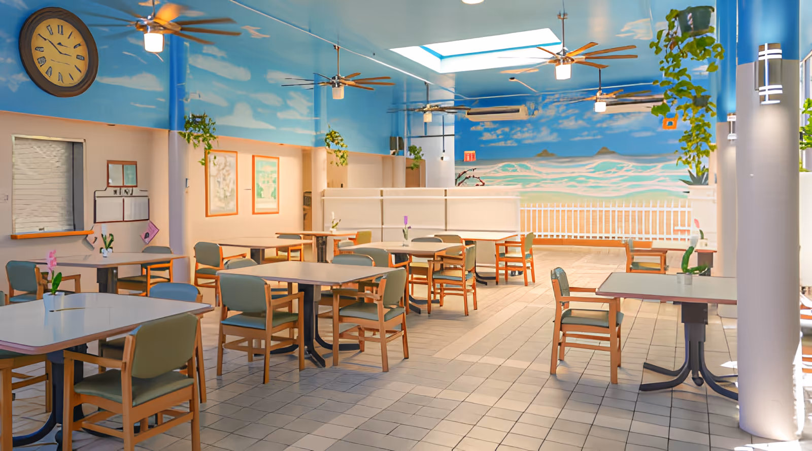 A bright dining room with several tables and chairs arranged neatly. The walls and ceiling are painted with a sky and clouds mural, and there are ceiling fans and hanging plants. A large clock is mounted on the wall, and there are framed pictures and a service window on one side. The room has tiled flooring and a skylight allowing natural light to enter.