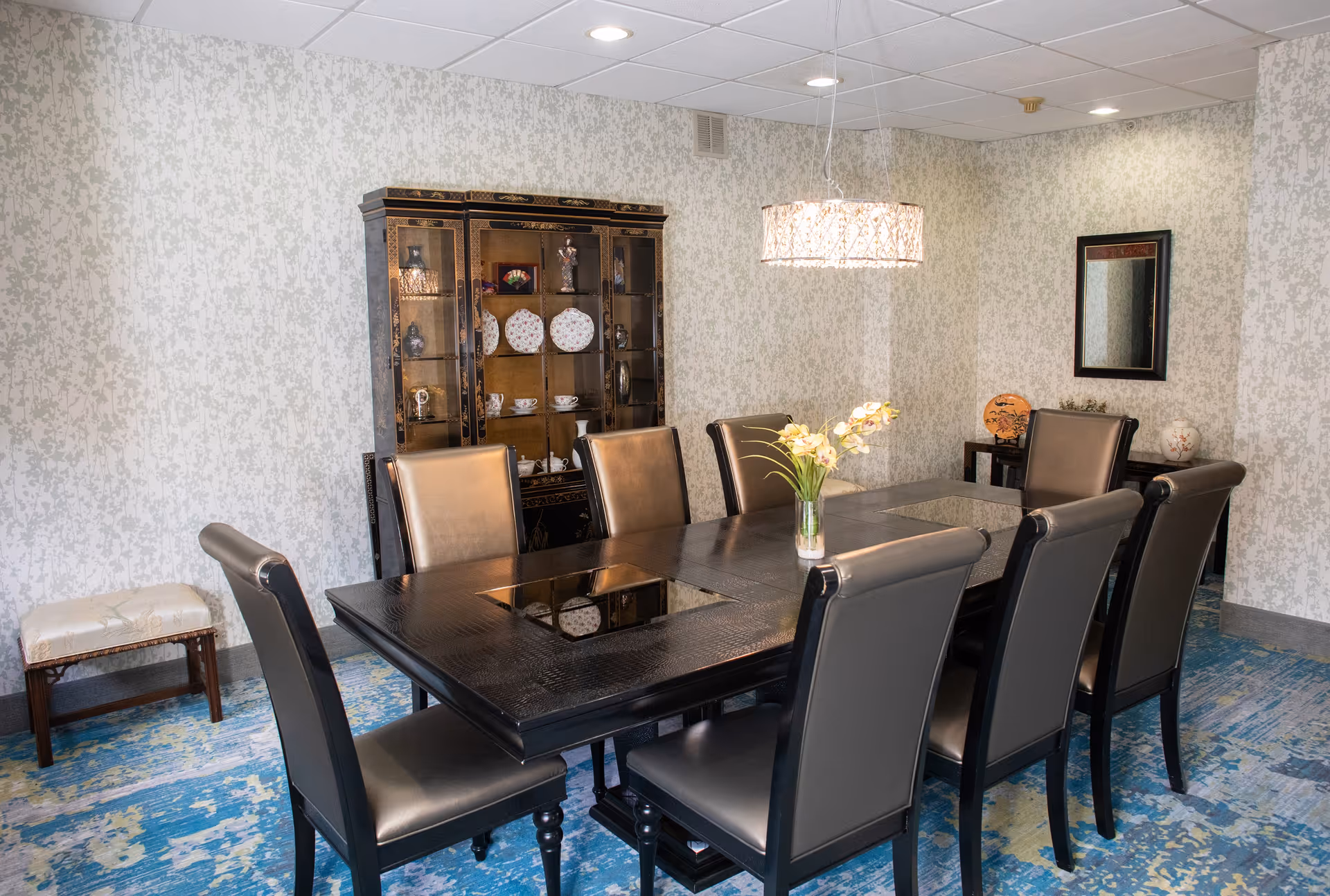 A dining room with a large rectangular dark wood table surrounded by eight black leather chairs. A glass vase with flowers is placed in the center of the table. Behind the table is a decorative cabinet displaying plates and teacups. The room has patterned wallpaper, a blue and yellow patterned carpet, a hanging light fixture above the table, a small bench on the left, and a mirror on the right wall above a small table with decorative items.