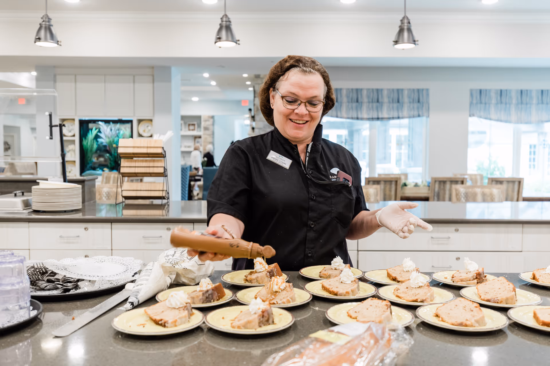 A woman wearing glasses and a black uniform with a name tag is smiling while pouring sauce on slices of dessert arranged on plates in a bright kitchen or dining area with white cabinets and large windows in the background.