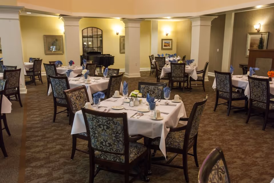 Spacious dining room with multiple tables set with white tablecloths, napkins, and upholstered chairs, and a piano in the background.