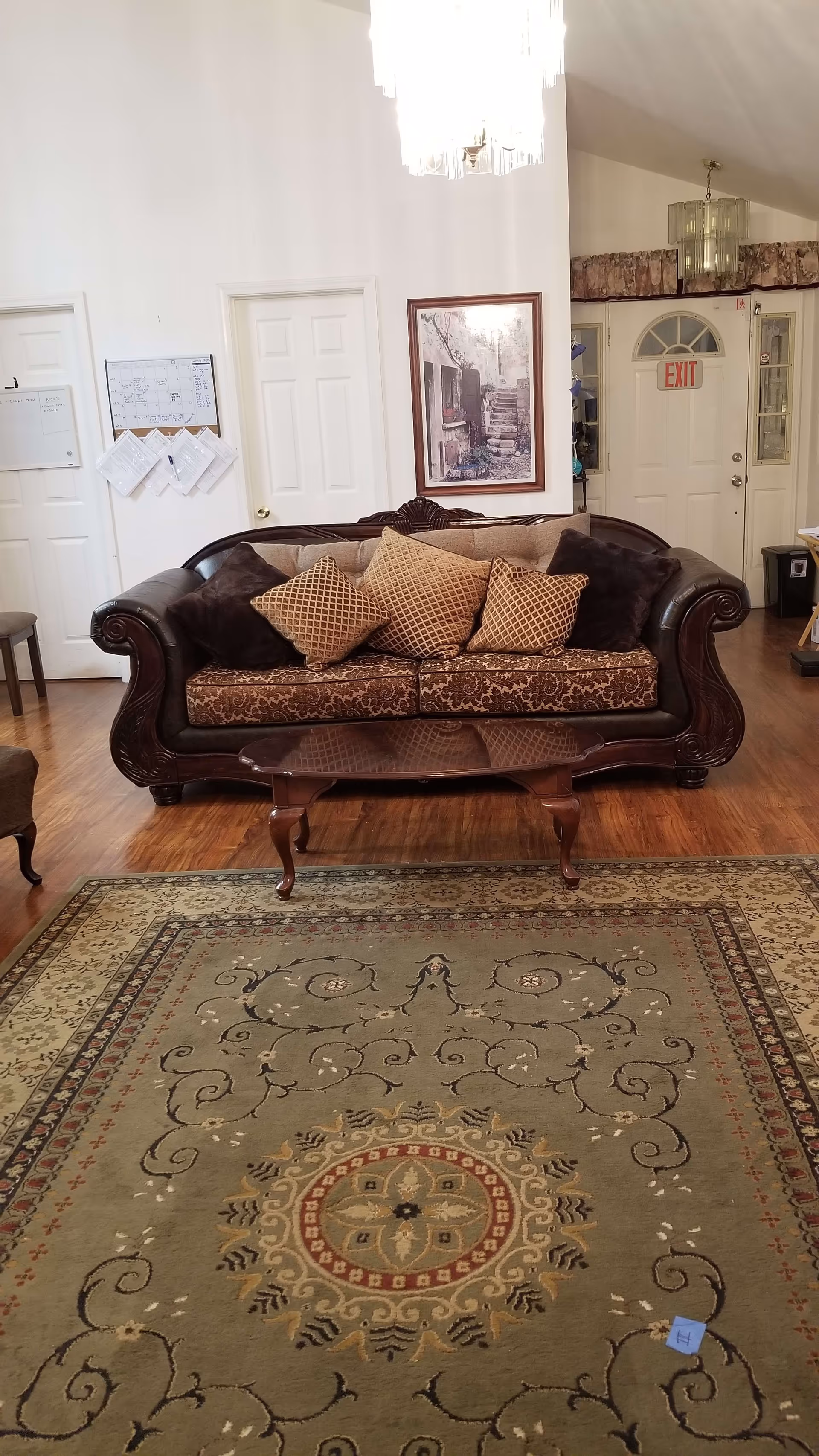 A living room with a dark brown ornate sofa adorned with five decorative pillows in shades of brown and beige. In front of the sofa is a wooden coffee table with curved legs. The floor features a large patterned area rug with intricate designs in green, beige, and red tones. The background shows white walls with two closed white doors, a framed picture of stone steps, an exit door with a red EXIT sign above it, and two chandeliers hanging from the ceiling.