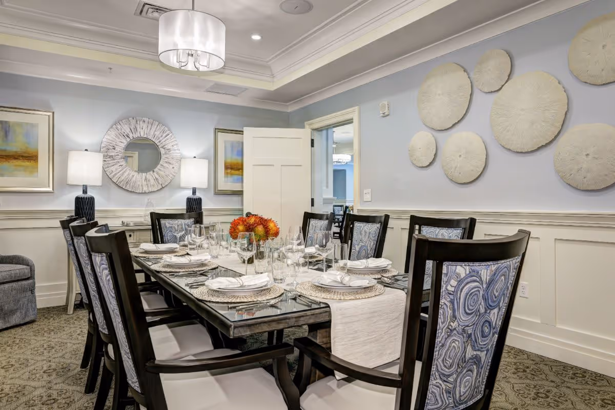 A well-decorated dining room with a long glass-top table set for eight people. The chairs have patterned blue and white upholstery. The table is set with plates, napkins, glasses, and a floral centerpiece. The room features light blue walls with white wainscoting, two framed paintings, a round decorative mirror, two table lamps, and circular wall art. A door is open leading to another room.
