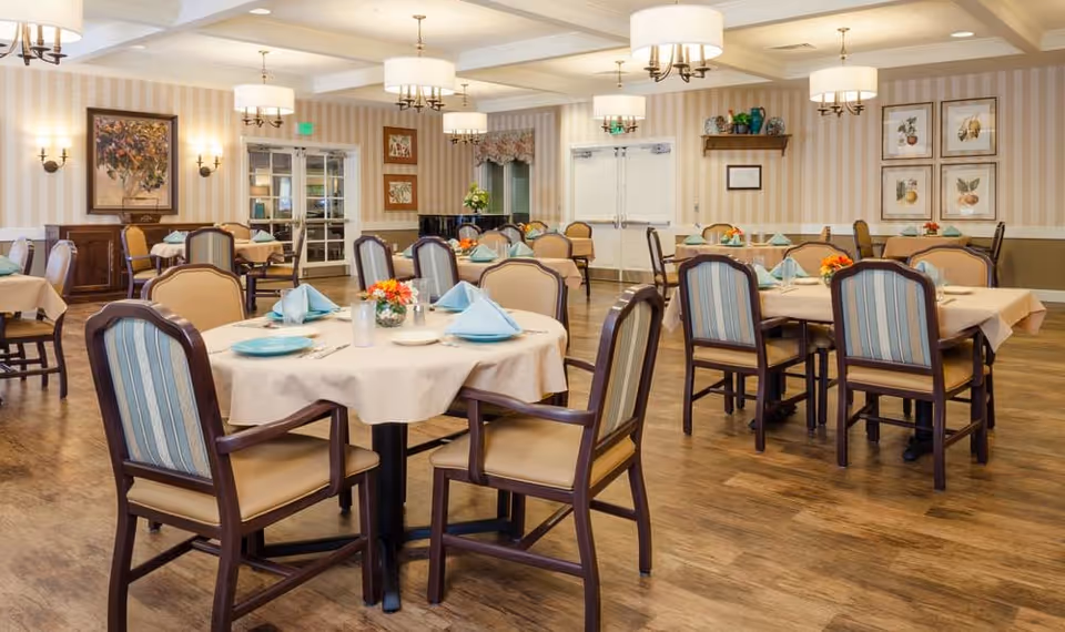 A spacious dining room with multiple round tables covered with beige tablecloths, each set with blue napkins, plates, and silverware. The room features wooden chairs with striped upholstery, hardwood floors, and soft lighting from ceiling fixtures and wall sconces. The walls are decorated with framed artwork and shelves with decorative items.