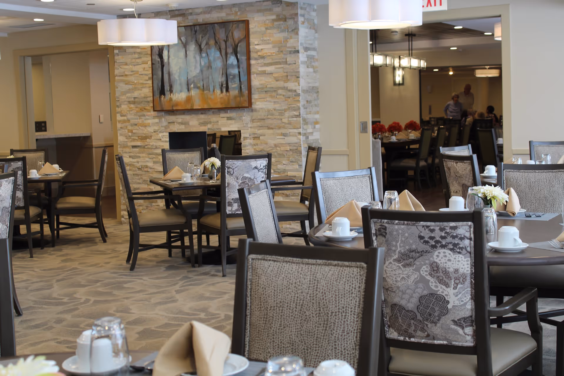 A dining room in a senior living facility with multiple tables and chairs arranged neatly. Each table is set with white cups, saucers, glasses, and beige napkins. The room features a stone accent wall with a framed painting of trees, soft lighting from ceiling fixtures, and a carpeted floor. In the background, a few people are visible near another dining area.