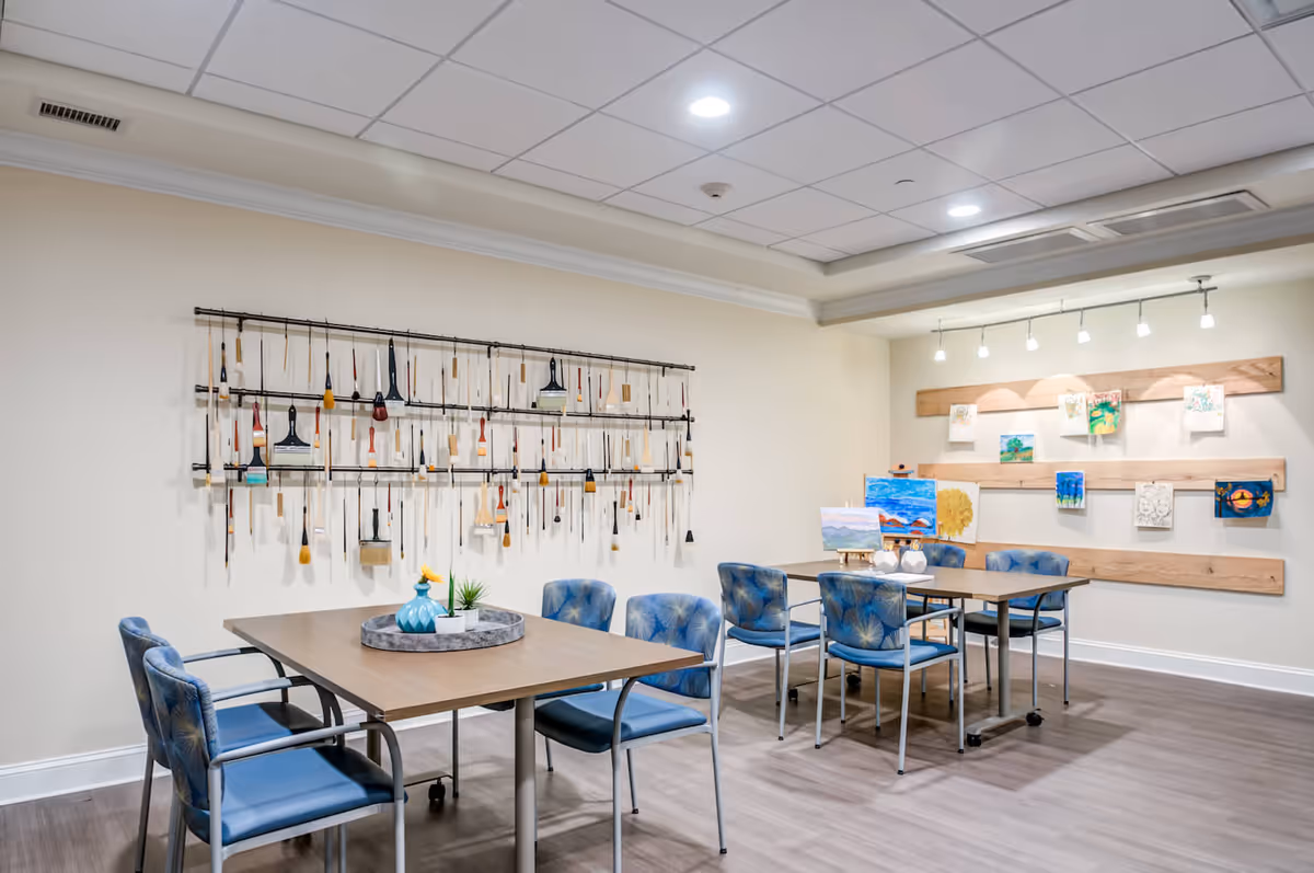 A bright art room with two tables surrounded by blue cushioned chairs. On the left wall, various paintbrushes are hanging on a metal rack. The right wall features wooden slats with several colorful paintings displayed. The room has a light-colored ceiling with recessed lighting and a wooden floor.