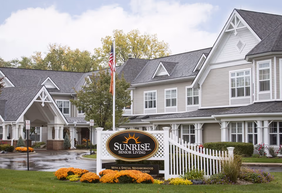 Exterior view of Sunrise Senior Living facility with a large sign in front, a white fence, and a well-maintained garden with orange and yellow flowers. The building has beige siding, white trim, and multiple windows under a dark gray roof. An American flag is flying on a flagpole near the entrance.