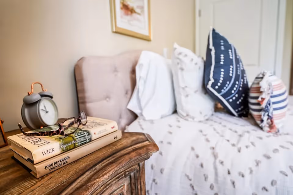 Close-up view of a bedside table with two books, a pair of eyeglasses, and a vintage-style alarm clock. In the background, a bed with a tufted headboard is partially visible, adorned with white and patterned pillows and a white bedspread with small designs.