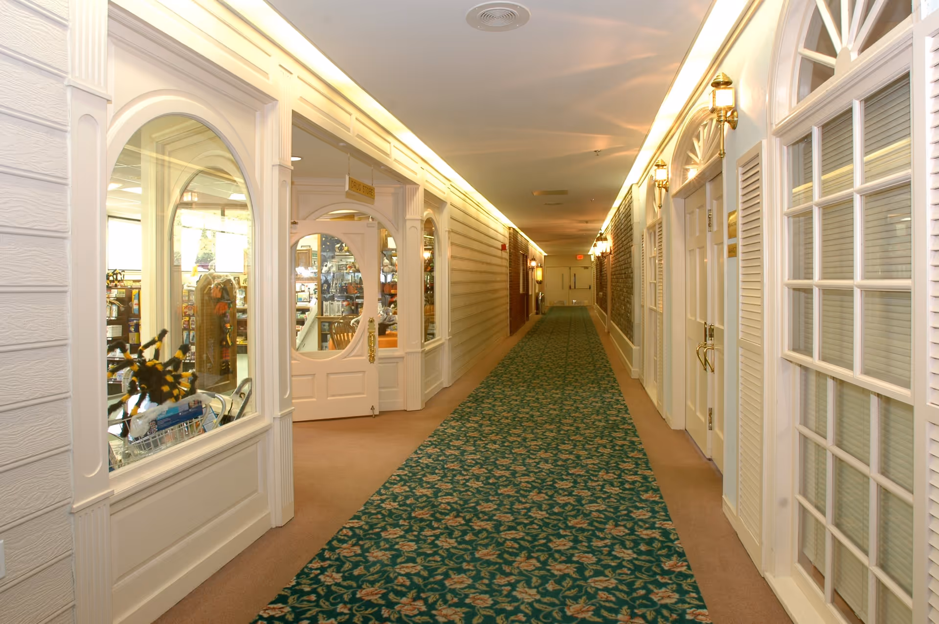 A long, well-lit indoor hallway with a patterned green carpet. On the left side, there is a storefront with large windows and a door labeled 'Drug Store,' displaying various items inside. The right side features white doors and windows with shutters, and the walls have decorative lighting fixtures. The hallway extends to double doors at the far end.