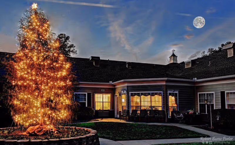 A large outdoor Christmas tree decorated with numerous bright white lights stands in the courtyard of a senior care facility at dusk. The building surrounding the courtyard has lit windows and a covered patio area with chairs. The sky is darkening with a visible full moon.