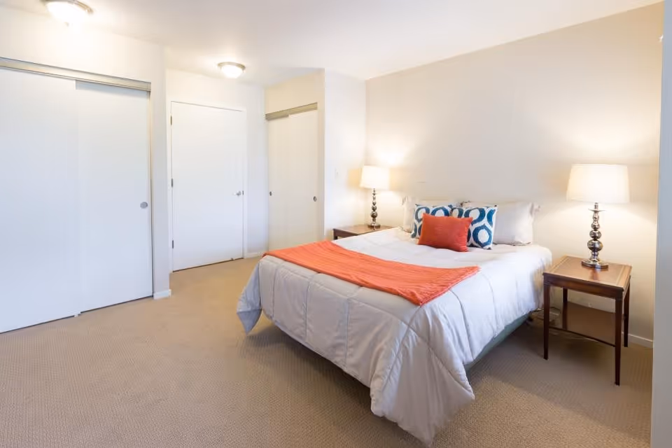A bright bedroom with a neatly made bed featuring white bedding, a red throw blanket, and decorative pillows. There are two bedside tables with matching lamps on either side of the bed. The room has beige carpet flooring, white walls, a closed white door, and two white sliding closet doors.