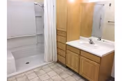 A bathroom featuring a walk-in shower with a white curtain, a wooden vanity with a white sink and faucet, a large mirror above the sink, and built-in wooden storage drawers and cabinets. The floor is covered with light-colored tiles.
