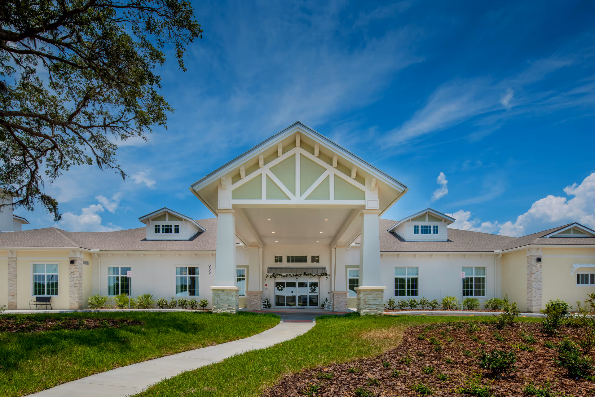 Front entrance of a single-story senior living building with a covered porte-cochere, manicured lawn, and blue sky.