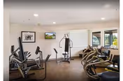 Interior view of a fitness room in a senior living facility with various exercise equipment including stationary bikes, elliptical machines, and weight machines. The room has large windows allowing natural light and framed pictures on the walls.