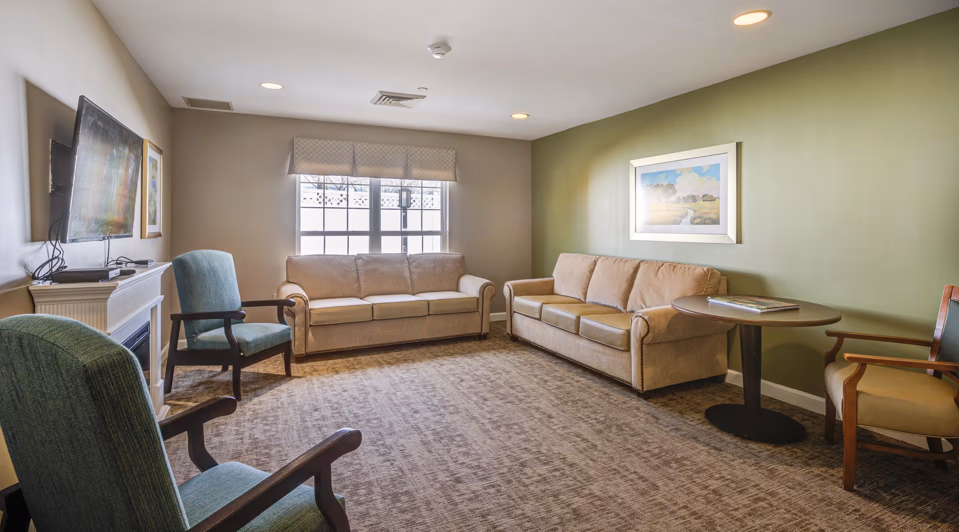 A cozy living room with two beige sofas, two green armchairs, a round wooden table with a book on it, a wall-mounted flat screen TV, a fireplace, and a large window with a valance. The walls are painted beige and green, and there is a framed landscape painting on the green wall.