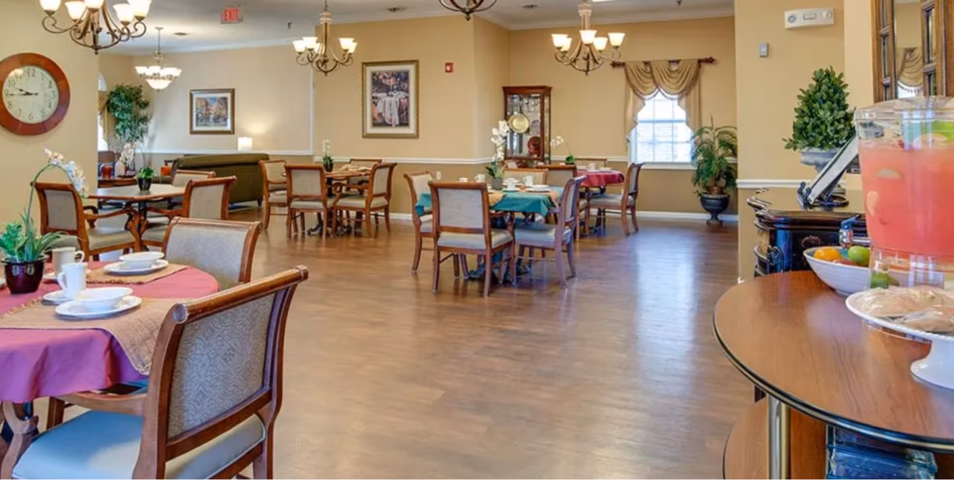 Bright senior dining room with multiple set tables and chairs, chandeliers, plants, and a refreshment table.