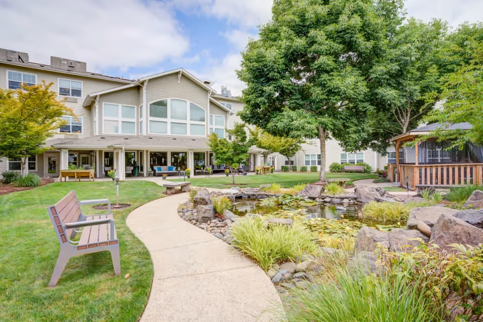 Outdoor garden area at Timber Pointe Senior Living featuring a curved concrete pathway, benches, a pond with rocks and water plants, large trees, and a multi-story building with large windows in the background.