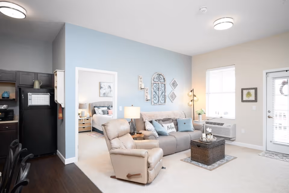 A bright and cozy living room with a beige recliner chair and a gray sofa adorned with decorative pillows. The wall behind the sofa is painted light blue and decorated with wall art. To the left, there is an open doorway leading to a bedroom with a bed and nightstand visible. The living room has a window with blinds, a floor lamp, and a small air conditioning unit below the window. A dark wicker coffee table sits on a small rug in front of the sofa. The kitchen area with dark cabinets and a black refrigerator is partially visible on the left side of the image.