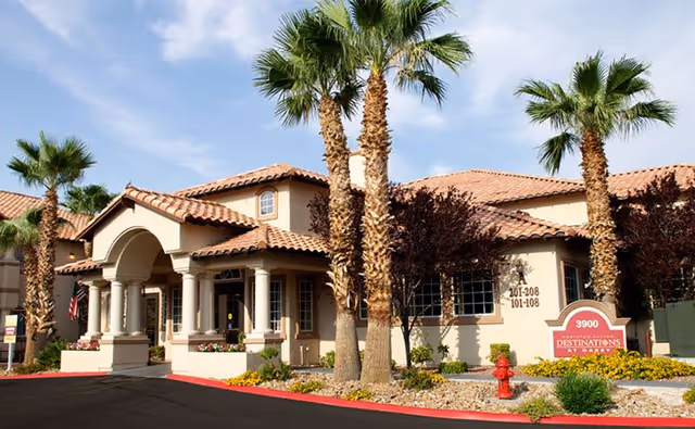 Exterior view of a single-story assisted living facility with a tiled roof, palm trees, and landscaped garden beds. The building has a covered entrance supported by columns and multiple windows. A red fire hydrant and a sign with the address 3900 and the name Destinations at Las Vegas are visible near the entrance.