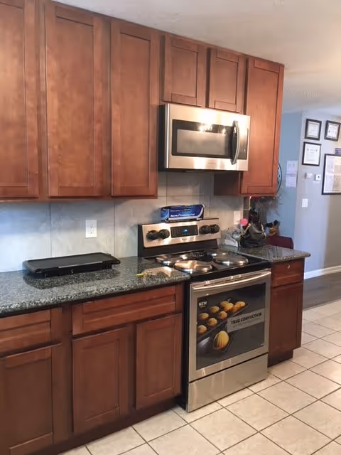 A kitchen interior featuring wooden cabinets, a granite countertop, a stainless steel microwave mounted above a stainless steel electric stove with four burners. There is a griddle on the countertop and some kitchen items near the wall. The floor is tiled, and part of a hallway with framed certificates on the wall is visible in the background.