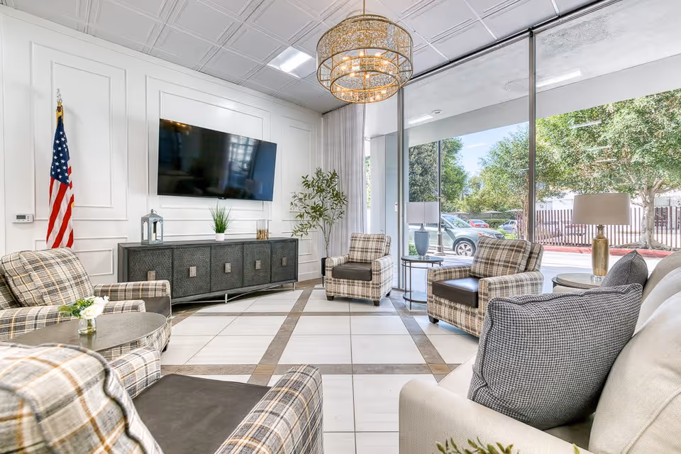 A bright and modern living room area with large windows showing an outdoor view. The room features plaid upholstered armchairs, a beige sofa with gray pillows, a round coffee table with a small flower arrangement, a large flat-screen TV mounted on a white paneled wall, an American flag, a decorative sideboard, and a large chandelier hanging from the ceiling.