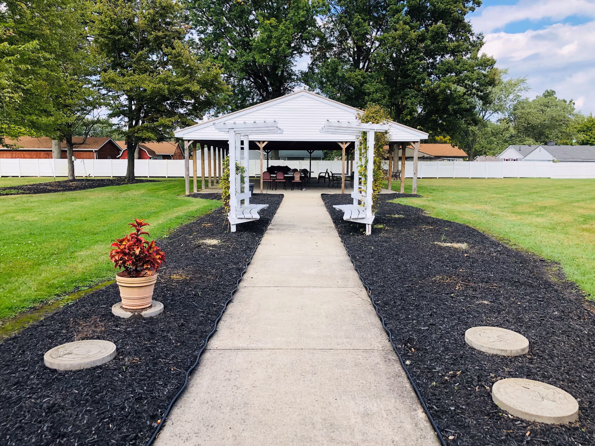A paved walkway leads to a white gazebo with benches and chairs underneath, surrounded by a well-maintained lawn and trees. There is a potted plant on the left side of the walkway and circular stepping stones embedded in the mulch on both sides. The sky is partly cloudy.