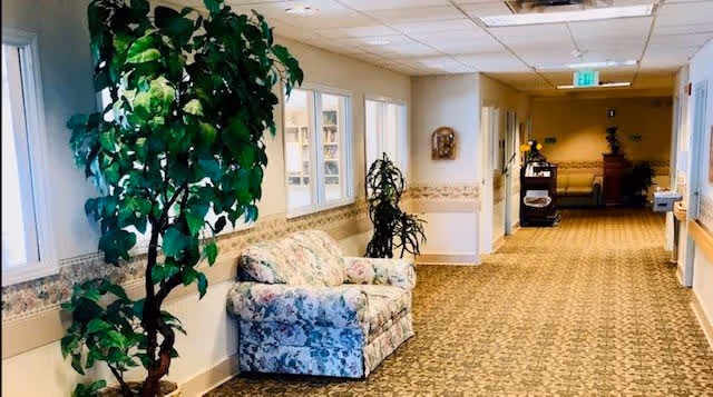 A long, carpeted hallway in a senior living facility with floral-patterned armchairs and potted plants along the walls. The walls are light-colored with a decorative border, and there are windows on the left side. The hallway is well-lit with ceiling lights and has an exit sign visible at the far end.