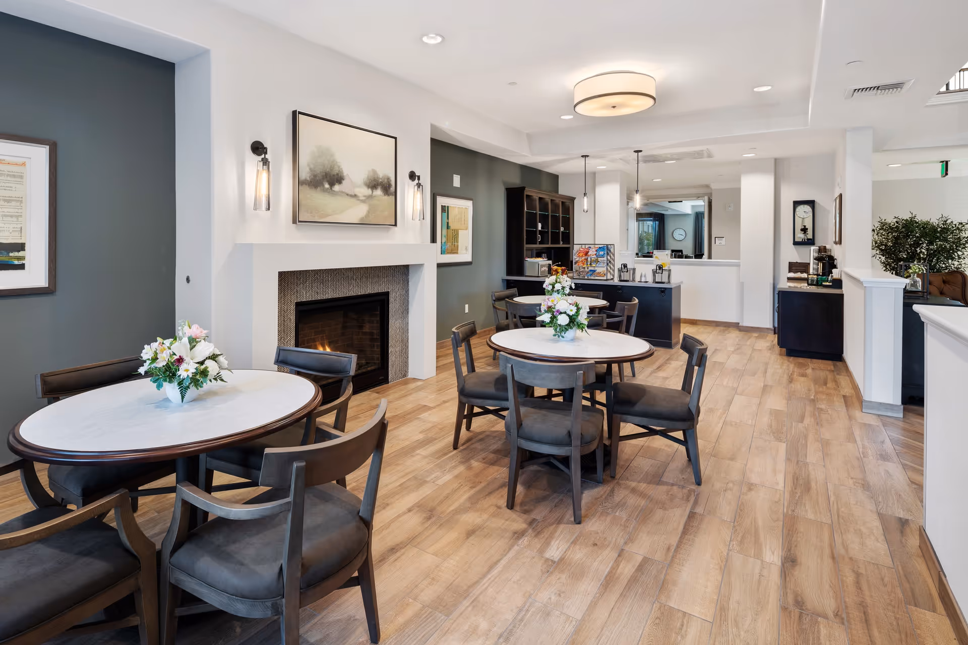 A cozy common area with round tables and chairs arranged on a wood floor. Each table has a floral centerpiece. There is a lit fireplace with a painting above it on the left wall. The room features modern lighting fixtures, framed artwork, and a small counter area with snacks and beverages in the background.