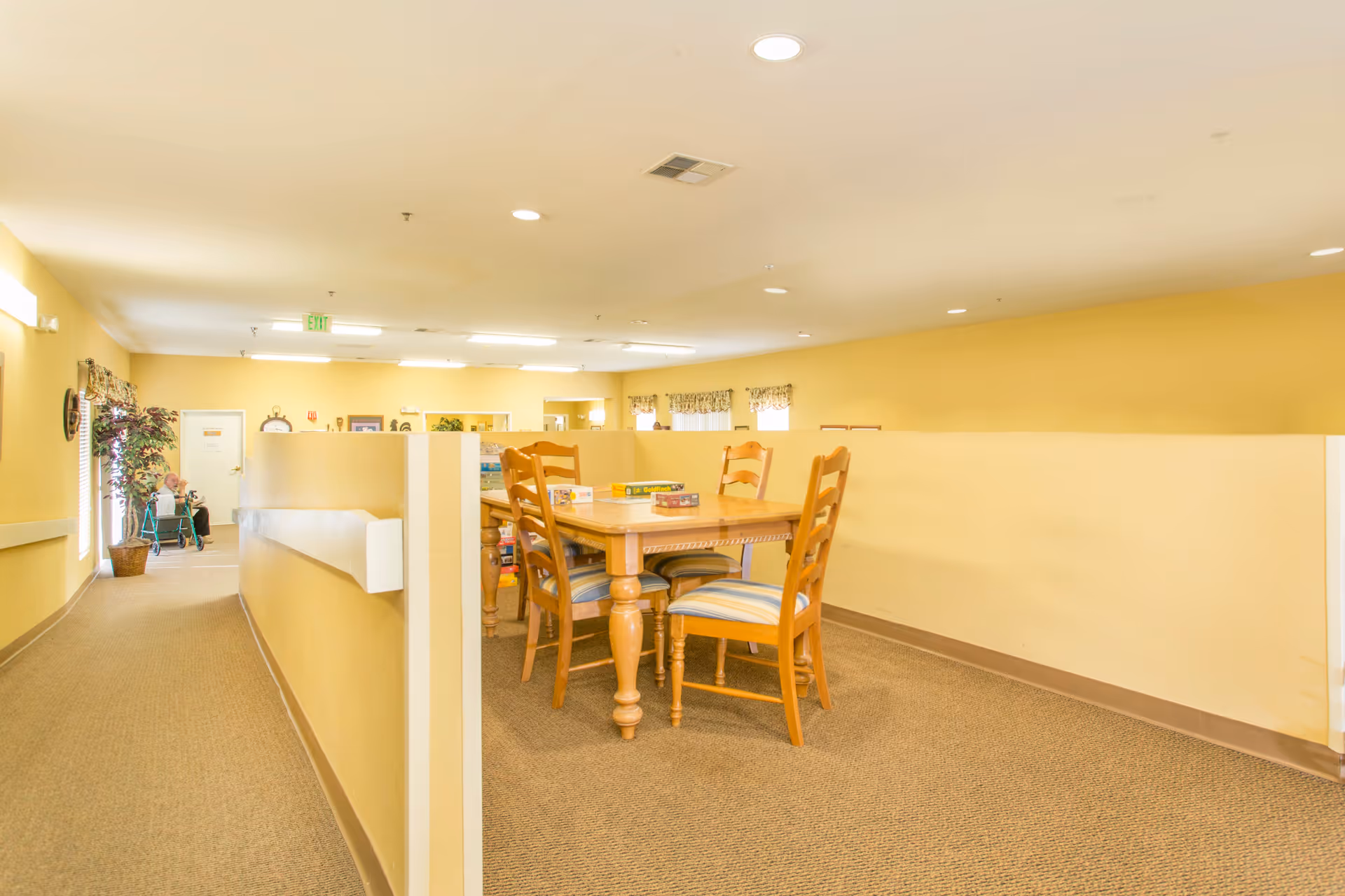 Interior of a senior living facility with yellow walls and carpeted floors. A wooden table with four chairs is placed in a semi-private area with low partitions. Board games are on the table. In the background, an elderly person with a walker is seated near a window with a potted plant beside them.