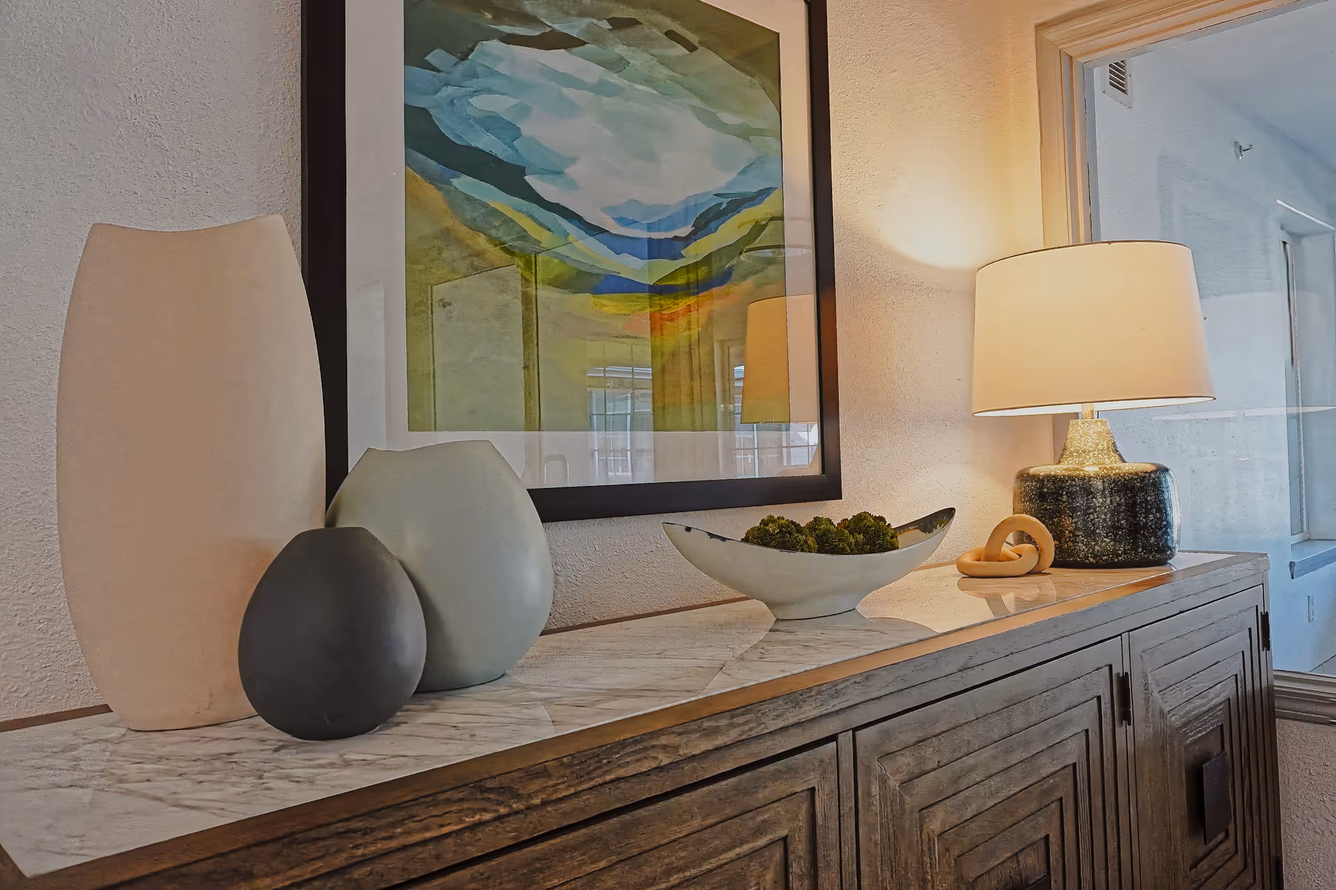 A wooden sideboard with a marble top displaying decorative items including three vases of different shapes and colors, a bowl with green moss balls, and a table lamp with a beige shade. Above the sideboard is a framed abstract painting with green, blue, and yellow tones. A window is visible to the right, showing an exterior wall.
