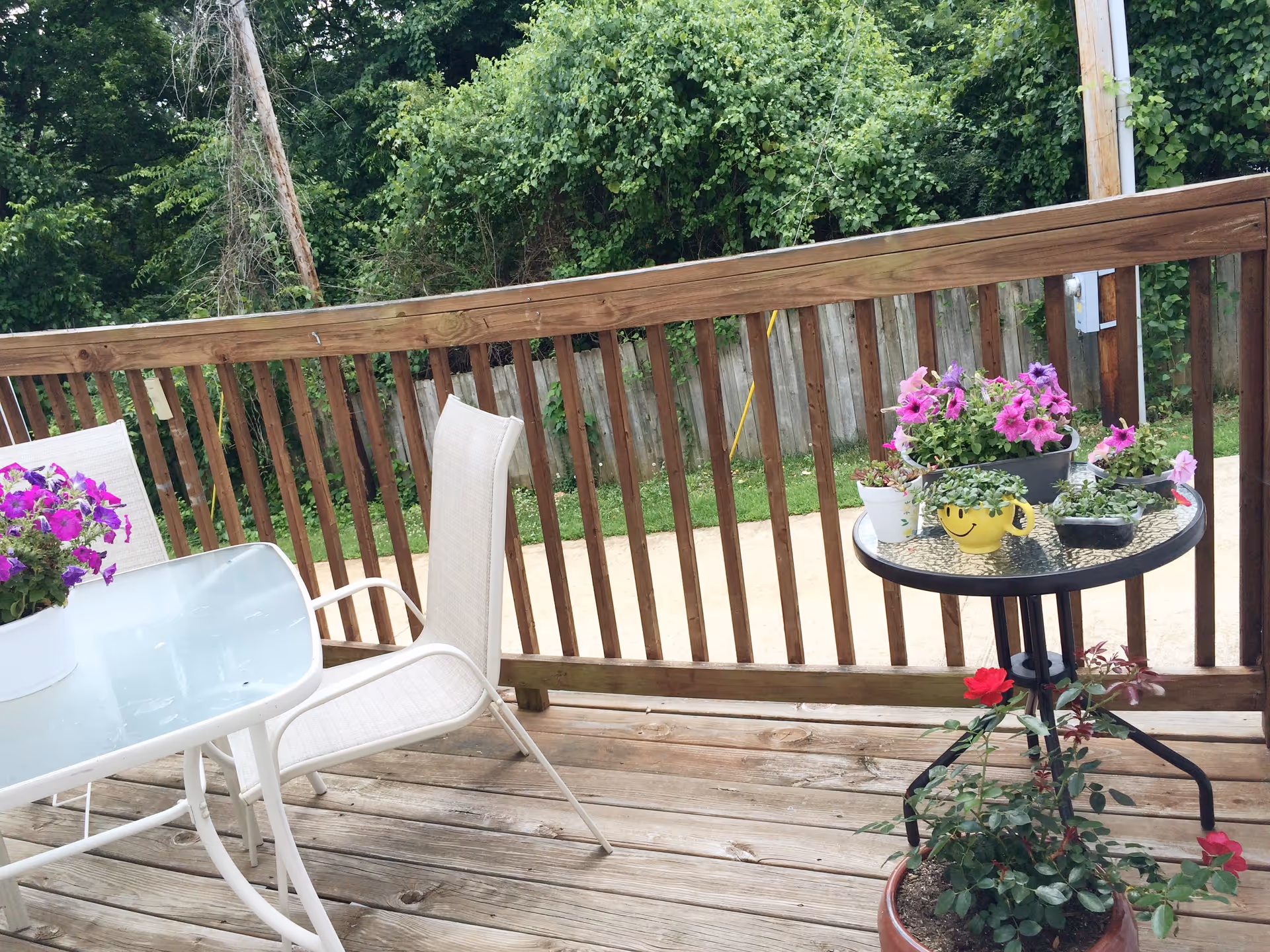 Outdoor wooden deck with a glass table and white chairs. Several potted plants with purple and red flowers are placed on the table and the deck. A wooden fence and green trees are visible in the background.