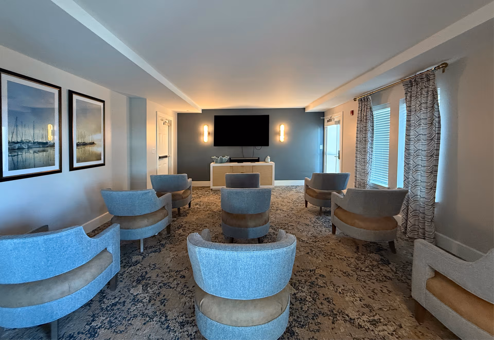 A cozy common room with multiple light blue and tan armchairs arranged in rows facing a wall-mounted flat screen TV. The room has a patterned carpet, two framed pictures of boats on the left wall, two wall sconces flanking the TV, and windows with patterned curtains on the right side.