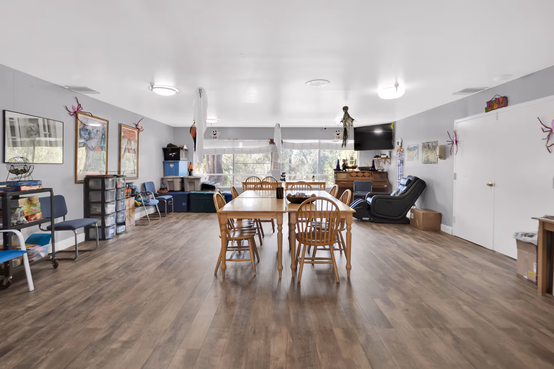 A spacious common room with wooden flooring featuring two wooden tables surrounded by chairs in the center. The room has large windows at the far end letting in natural light, a TV mounted on the wall, a recliner chair, and various storage bins and chairs along the walls. The walls are decorated with framed pictures and Halloween-themed decorations.