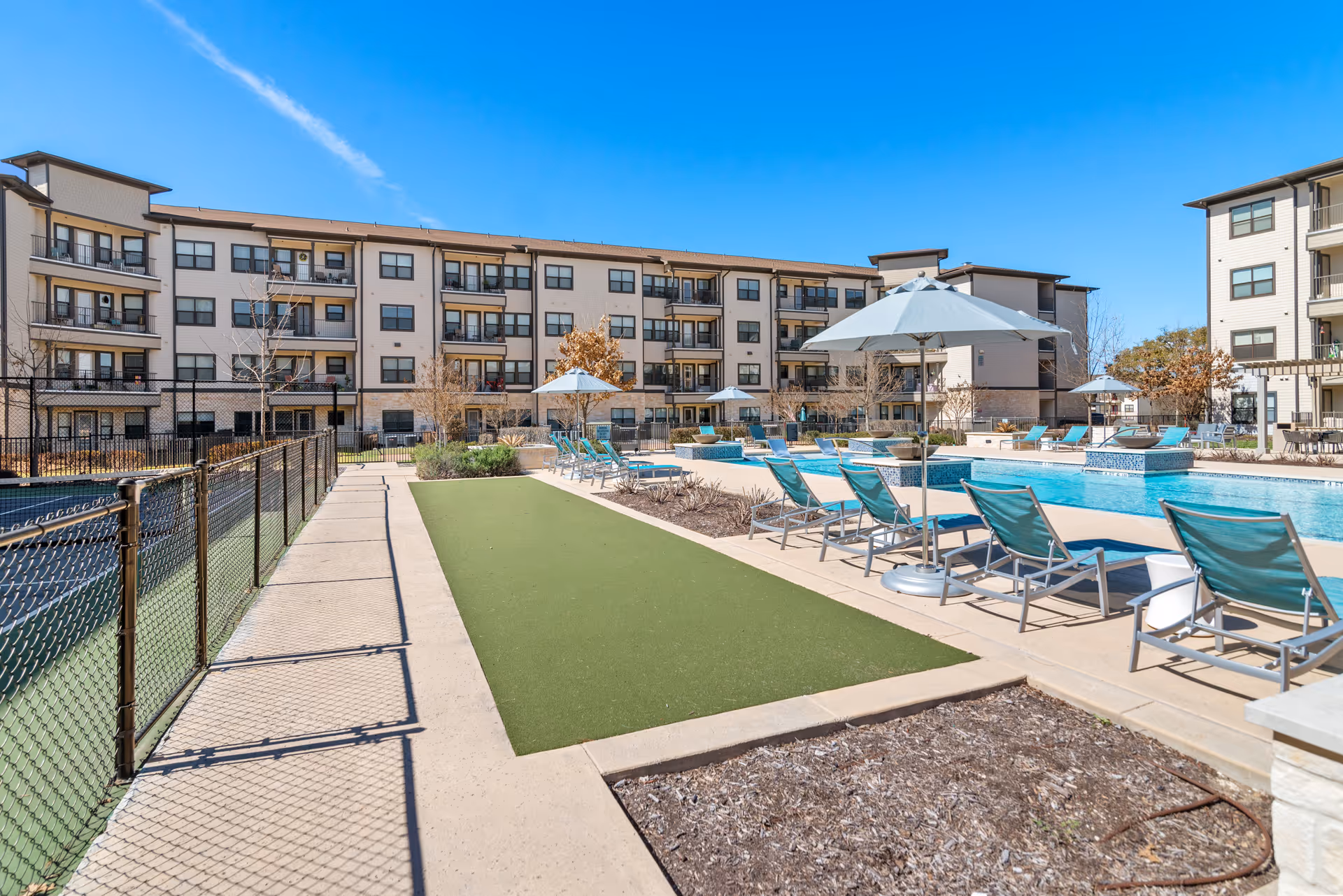 Outdoor area of Solea Alamo Ranch featuring a swimming pool with blue lounge chairs and umbrellas, a bocce ball court, and a multi-story residential building in the background under a clear blue sky.