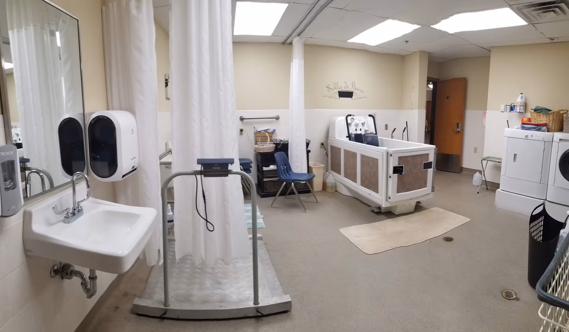 Accessible bathing room in a care facility with a sink, shower curtains, a patient lift platform and a wheelchair-accessible bathtub.