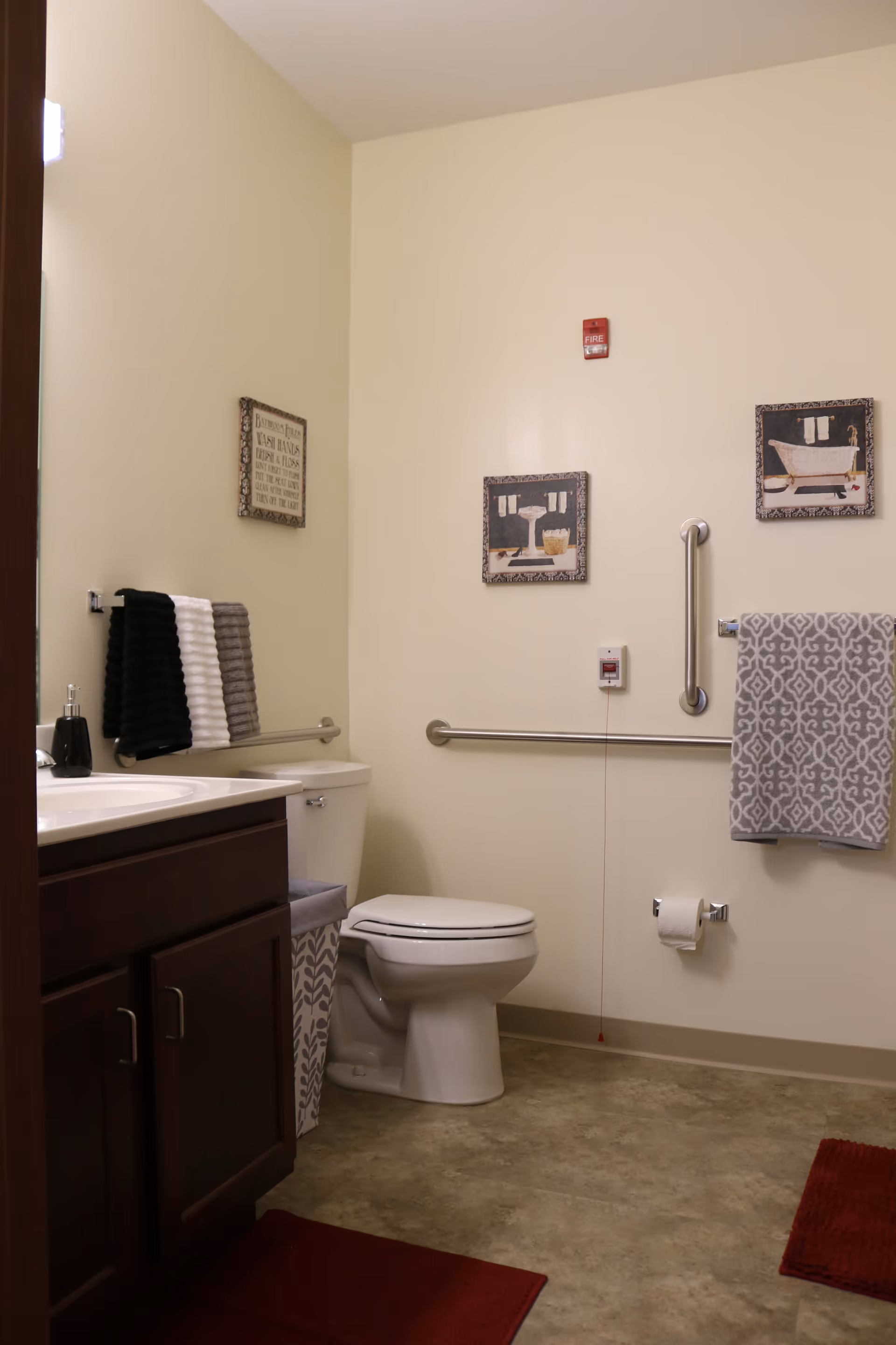 A clean and accessible bathroom with a white toilet, dark wood vanity with a sink, and grab bars on the walls. There are decorative framed pictures on the wall, a towel rack with a gray patterned towel, and a set of black, white, and gray towels hanging near the sink. The floor has a beige tile pattern with red bath mats.