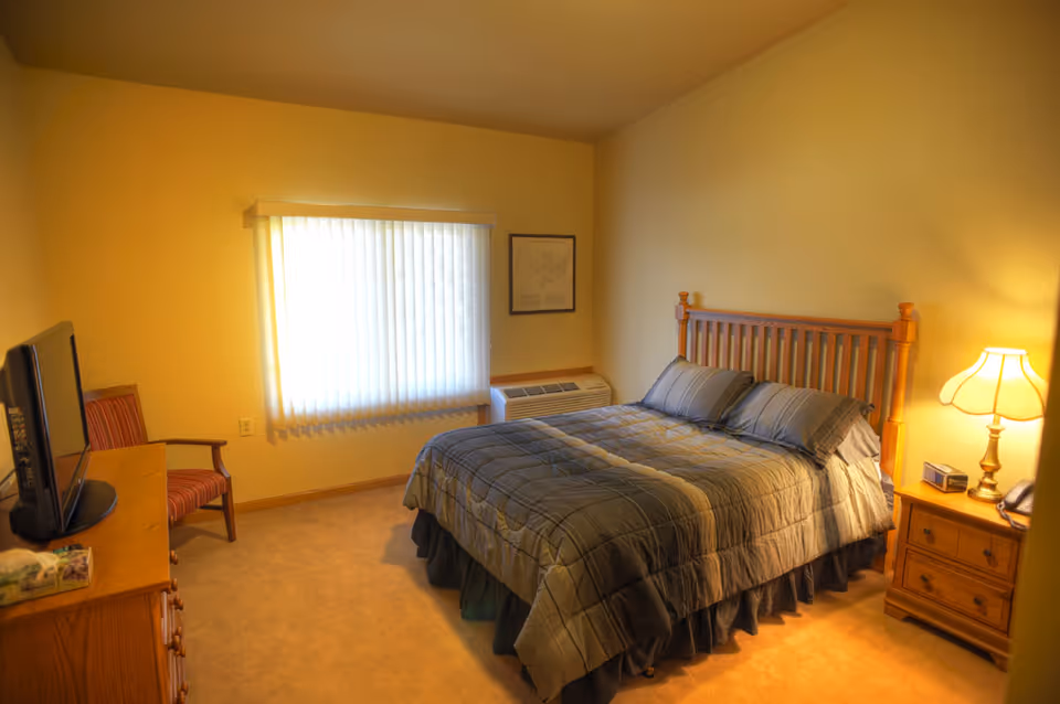 Well-lit bedroom with a made bed featuring a wooden headboard, bedside table and lamp, a TV on a dresser, and a window with vertical blinds.