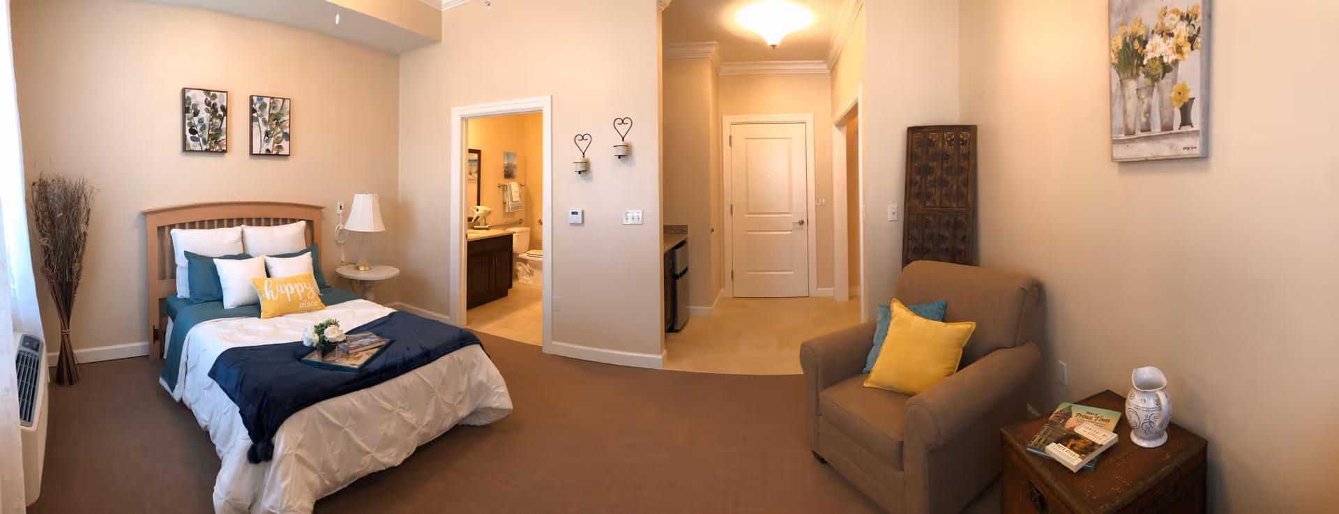 A cozy senior living bedroom with a neatly made bed featuring white and teal bedding and a yellow pillow that says 'happy'. There is a small round side table with a lamp next to the bed, and a tall vase with decorative branches in the corner. The room has beige walls with two framed botanical prints above the bed. To the right, there is a comfortable armchair with yellow and teal pillows, next to a wooden side table holding books and a decorative white vase. A doorway leads to a bathroom with visible sink and toilet, and another doorway leads to a hallway with a closed white door. The room is well-lit with natural light coming from a window on the left.