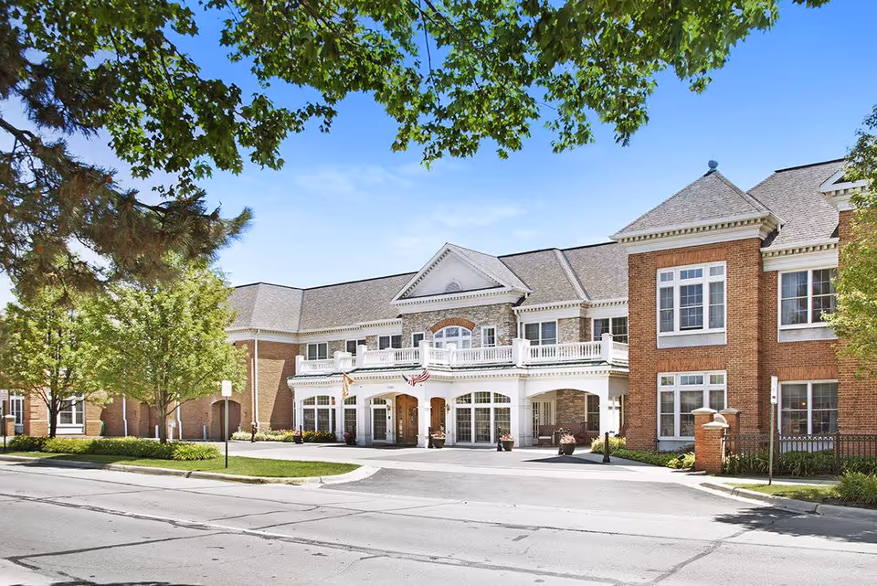 Entrance and facade of a two-story brick senior living building with a covered porte-cochère, balcony, American flag, and surrounding trees.